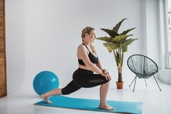 A plus size woman in a black sports top and leggings is doing morning exercises on a yoga mat. Chubby blonde is engaged in fitness. Curvy girl doing stretching in a bright room sitting on the floor