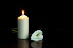 WHITE CALLA FLOWER NEXT TO A LIGHTED CANDLE ON DARK BACKGROUND. ALL SOULS DAY, DEATH, DECEASE, PRAYER, MEMORIAL DAY, MOURNING, GRAVE, CEMETERY, BURIAL, CONDOLENCE AND FUNERAL CONCEPT. COPY SPACE.