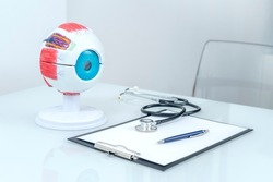 OPHTHALMOLOGIST'S DESK IN THE OFFICE WITH HUMAN EYE ANATOMY MODEL, STETHOSCOPE, PEN AND NOTEBOOK.
