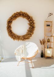 A wicker round chair stands against the wall, a white soft plaid on the chair. Beautiful shadows and sunlight. Bright and spacious room. On the wall is a decoration made of dry reeds and grass.