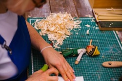 A wooden toy in women's hands. A female carpenter cuts wood with a knife in a carpentry or makeshift workshop. Dust and shavings fly in the air.Workplace.