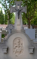 selective focus of a tombstone of a Catholic burial with a cross and a bronze sculpture of Christ and a relief of the Virgin of Carmen worn by time under the shade of some trees