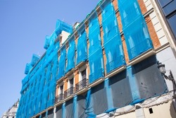 Reconstruction of facade of a historic classical apartment building on a city street in sunny day against sky. Blue facade construction mesh covers an old red house closed for renovation wall exterior