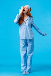 What wonderful morning. Full-length vertical studio shot yawning cute caucasian redhead girl in sleep mask and pyjama, stretching and cover opened mouth with closed eyes, blue background