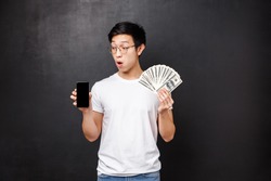 Technology, money and prizes concept. Amazed and surprised young asian guy still cant believe he won money via giveaway in internet, showing mobile phone display and dollars cash