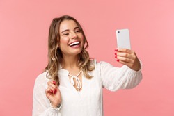 Close-up portrait of cheerful upbeat smiling blond girl, wearing white dress, laughing as record video, calling friend on mobile application, taking photo, selfie on smartphone, pink background