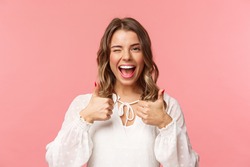 Portrait of sassy good-looking blond girl assure all good, show thumbs-up and wink with classy pleased smile, recommend visit place, shop at online store, standing in white dress pink background