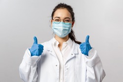 Doctors, infectionist, research and covid19 concept. Satisfied young asian female doctor receive good results on studying virus, discover vaccine, show thumb-up, wear face mask and gloves