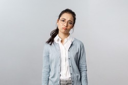 Portrait of sighing displeased and gloomy young asian girl in glasses, pouting and looking disappointed camera, express regret or feel uneasy over failure at work, grey background