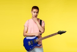 Lifestyle, leisure and youth concept. Unsure cute asian guy playing in band, thinking, making decision during band discussion what play, hold electric guitar, touch chin pondering at camera