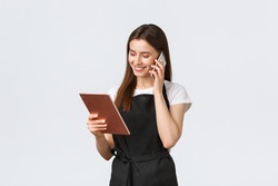 Grocery store employees, small business and coffee shops concept. Cute smiling saleswoman in black apron confirm order, talking to customer on phone and looking at digital tablet