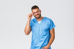 Healthcare workers, medicine, covid-19 and pandemic self-quarantine concept. Handsome cheerful male doctor, nurse in blue scrubs and glasses, smiling camera friendly and happy