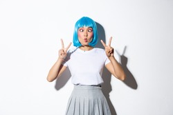 Portrait of silly and cute asian girl in blue wig, squinting and making funny faces, showing peace gestures, standing over white background