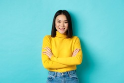 Confident and stylish asian woman cross arms on chest and smiling, standing over blue background