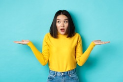 Confused and shocked asian woman dont understand, shrugging with spread hands, gasping and looking left, know nothing, standing over blue background
