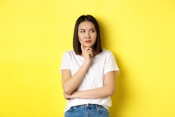 Beauty and fashion concept. Pensive asian woman thinking, looking thoughtful while pondering something, standing over yellow background