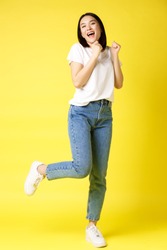 Full size shot of happy asian woman dancing and jumping from happiness, winning and celebrating victory, posing over yellow background