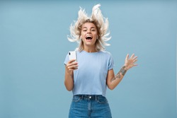 Expressing happiness with help of great tunes. Joyful amused and happy good-looking young female student jumping having fun listenign music in wireless earbuds, holding smartphone over blue wall