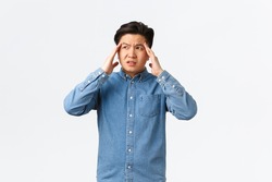 Troubled and uneasy asian man looking perplexed, having complicated situation. Guy with headache touching head and looking away, suffering painful migraine, standing white background