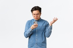 Frustrated and confused asian guy in glasses looking puzzled at mobile phone screen, raising hand up questioned, cant understand what happening online, standing white background perplexed