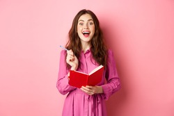 Excited young woman have excellent idea, writing down her ideas in planner, holding notebook diary and smiling amazed, standing over pink background