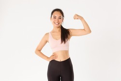 Sport, wellbeing and active lifestyle concept. Portrait of smiling slim and strong asian fitness girl, personal workout trainer showing muscles, flexing biceps and look proud, white background
