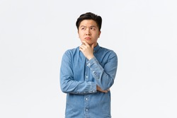 Thoughtful complicated asian man in shirt, touching chin and looking upper left corner, thinking, making decision, choosing something, having doubts, standing hesitant over white background