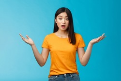 Questioned unaware young asian girl with dark short haircut shrugging hands spread sideways, look confused open mouth uncertain, being clueless, stand blue background wear yellow t-shirt