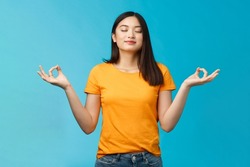 Peaceful charming relaxed asian girl buddhist meditating, breathing, inhale fresh air practice yoga, close eyes smiling relieved, stand lotus nirvana pose, reach zen, standing blue background
