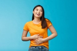 Happy staffed cute asian girl feelings satisfaction after eating favorite food, smiling broadly rubbing tummy, touch stomach from delight and joy, like eat delicious meals, stand blue background