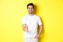 Handsome and confident man extending hand for handshake, greeting you, saying hello, standing in white t-shirt over yellow background