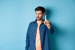 Image of young man give warning, teaching a lesson, raising one finger to scold, looking at camera, patronizing someone, standing on blue background