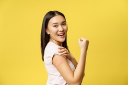 Covid-19 and healthcare medical concept. Cheerful asian girl shows arm with patch after coronavirus vaccination, getting shot of vaccine, standing happy over yellow background