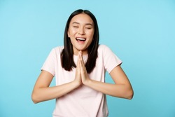 Smiling happy asian woman shows namaste, beg gesture and dreaming of something, making wish, high hopes, standing over blue background