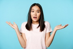 Confused asian girl spread empty hands sideways, shrugging shoulders clueless, standing in tshirt over blue background