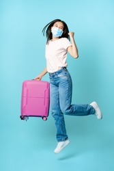 Full length of happy asia female tourist, dancing and jumping with suitcase in medical face mask, excited about vacation, standing over blue background