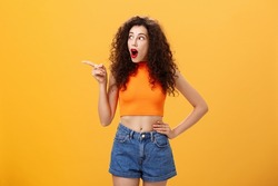 Woman peeking at neighbours window being surprised and amazed pointing and staring speechless left standing over orange background with dropped jaw intrigued and thrilled look in cropped top