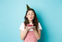Holidays and celebration. Excited woman celebrating birthday, blowing candle on cake, wearing party cake and having fun, standing over blue background