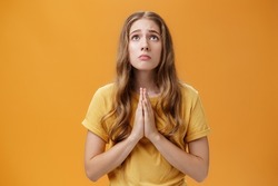 Hopeful uneasy and worried gloomy faithful girl in t-shirt holding hands in pray against chest looking up with sad look praying making wish for good well of family posing over orange wall