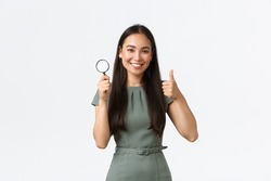 Small business owners, women entrepreneurs concept. Satisfied asian woman searching for something, showing thumbs-up after finding it, holding magnifying glass and smiling pleased, white background