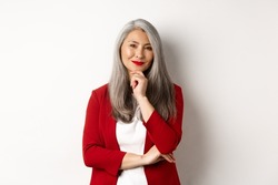Business concept. Asian mature businesswoman smiling pleased, looking thoughtful, having an idea, standing in red blazer over white background