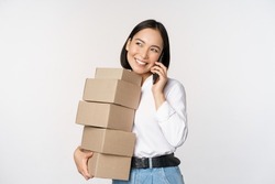Image of young asian businesswoman answer phone call while carrying boxes for delivery, posing against white background
