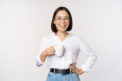 Happy young energetic asian woman smiling, drinking, holding cup mug of coffee, standing confident against white background