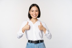 Image of beautiful adult asian woman showing thumbs up, wearing formal office, university clothing, recommending company, standing over white background