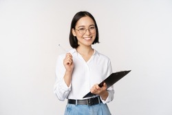 Smiling young asian woman taking notes with pen on clipboard, looking happy, standing against white background