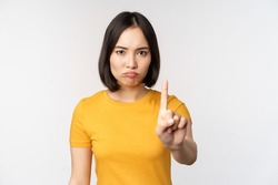 Portrait of asian woman looking serious and angry, showing stop prohibit gesture, taboo sign, forbidding smth, standing in yellow tshirt over white background
