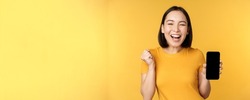 Excited asian woman, showing smartphone app and triumphing, celebrating on mobile phone, standing over yellow background