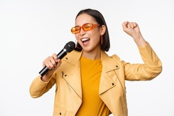 Stylish asian girl in sunglasses, singing songs with microphone, holding mic and dancing at karaoke, posing against white background