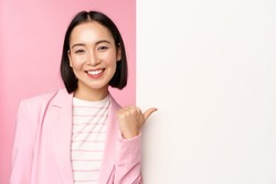 Portrait of smiling asian businesswoman in suit, corporate lady pointing finger at white empty wall, board with info or advertisement, standing over pink background