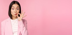 Portrait of asian corporate woman showing mouth seal, close shut lips on key gesture, promise keep secret, standing over pink background in suit